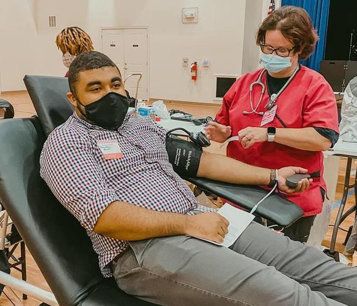 black male in plaid shirt and grey chinos giving blood wearing a face covering