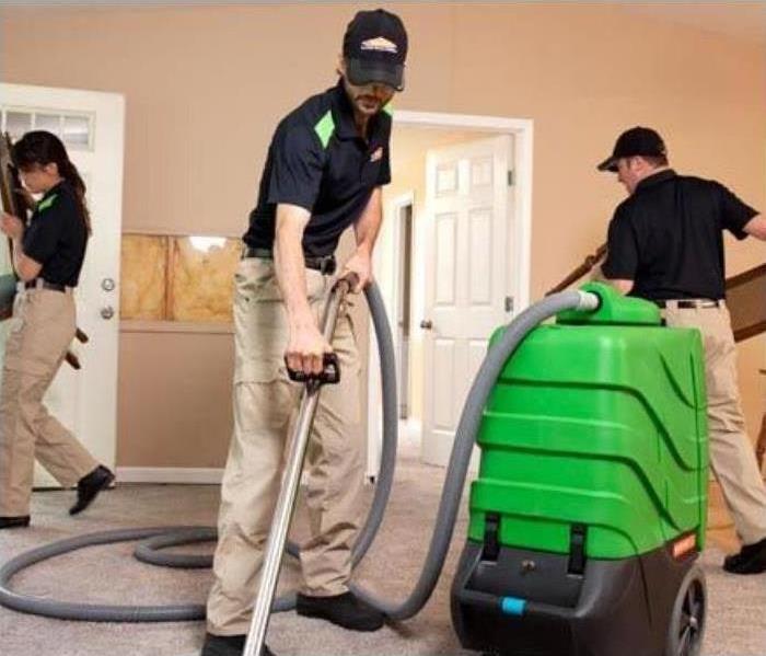 SERVPRO employees in company polo and khakis, wearing company hats, cleaning tan carpet with vacuum. door open on the left.