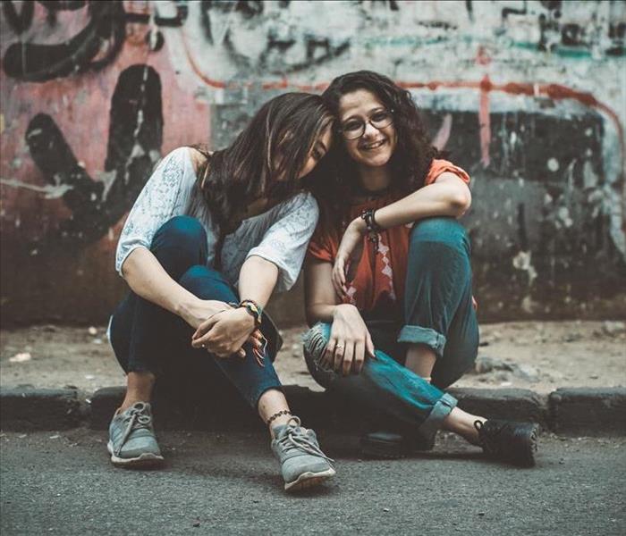 Two teen girls sit on a curb, hugging and smiling